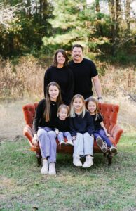 Family of six smiling outdoors on a red sofa with trees in the background.