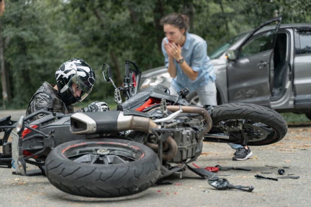 Motorcycle accident scene with fallen bike and rider on road while a concerned woman stands nearby