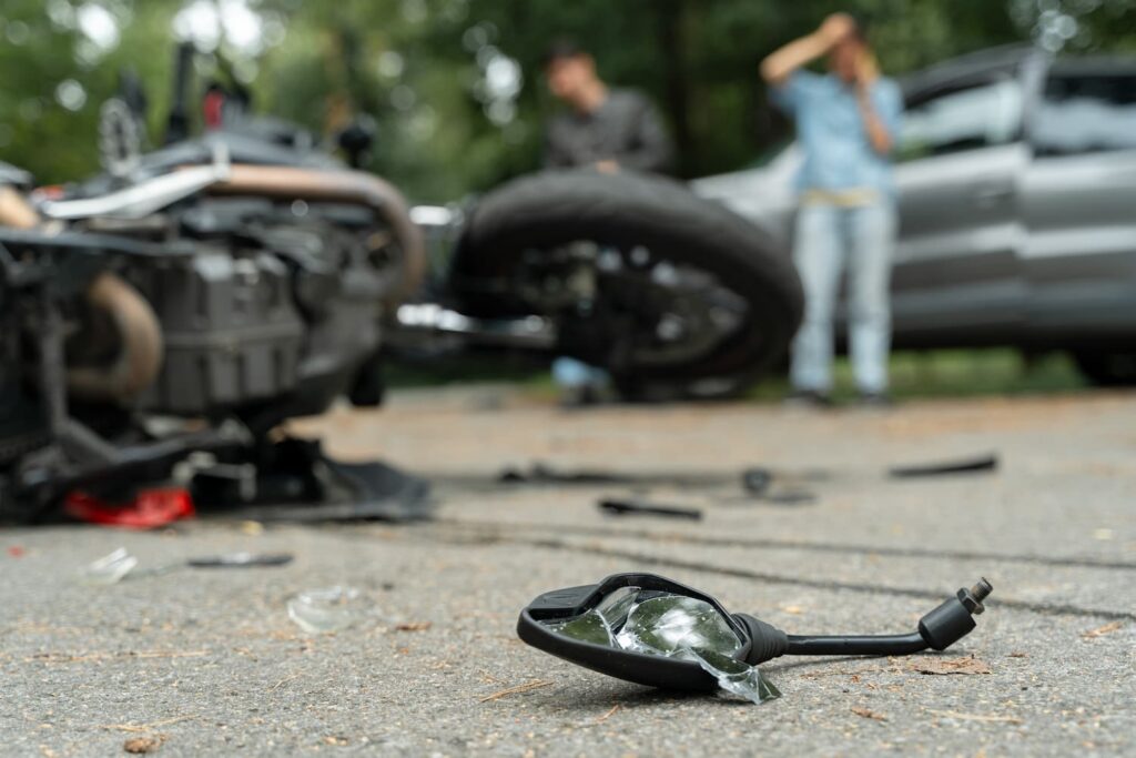 Motorcycle accident scene with damaged bike and broken side mirror on road, car and people in background